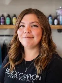 Smiling person in a black Canyon Performance T-shirt, standing in front of shelves of colorful bottles.