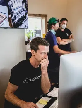 Three people working at computers in an office, one man in black focused on a monitor.