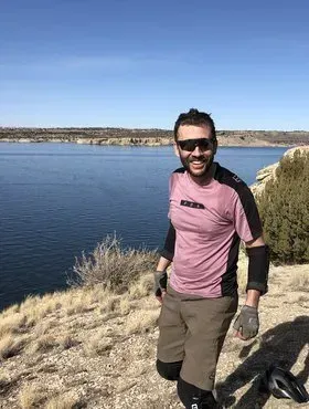 Man standing by a lake on a sunny day, wearing sunglasses and a pink shirt, with dry grass and shoreline behind him.