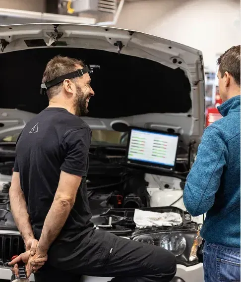 Two people examining a laptop in a car with the hood open, in a workshop