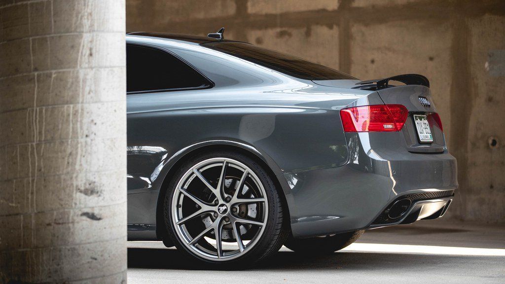 Gray sports car parked beside a concrete wall, rear three-quarter view with custom black wheels