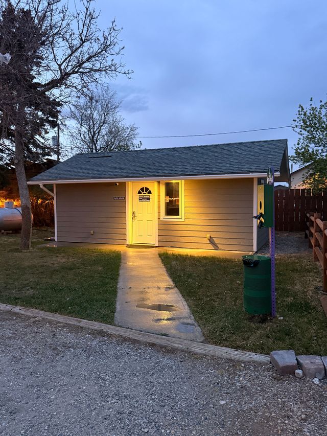 Small beige house at dusk with a lit front door and walkway, beside a gravel drive and grassy yard