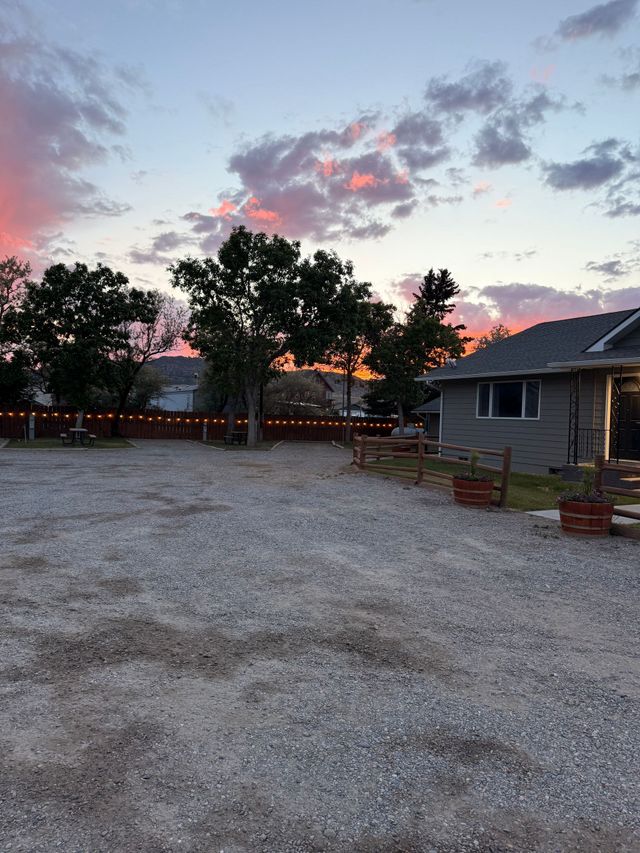 Gravel courtyard at dusk with trees, benches, potted plants, and a small house under a pink-blue sky