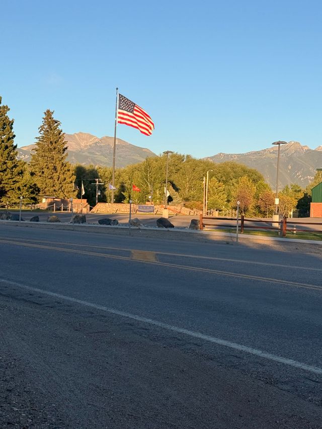 Large American flag by a roadside with trees and mountains under a clear blue sky at sunset