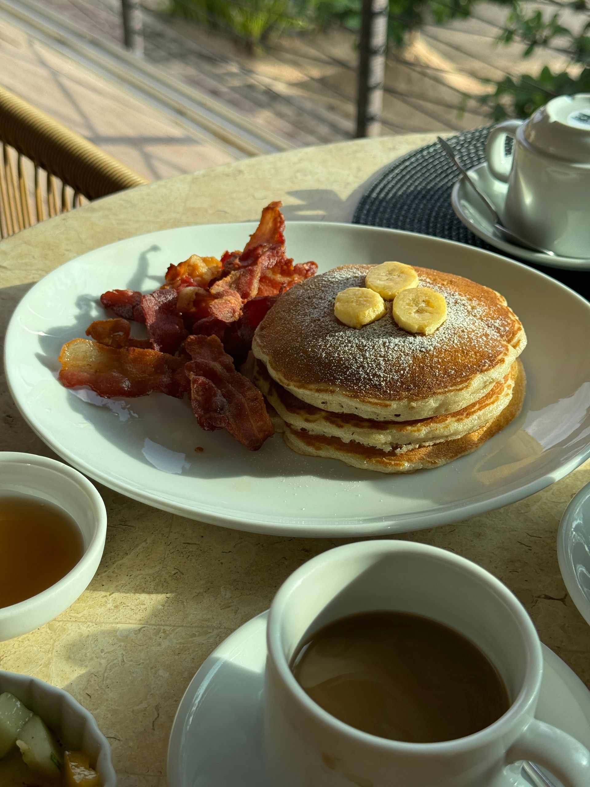 Pancakes with butter and bacon on a white plate beside two cups of coffee on a café table