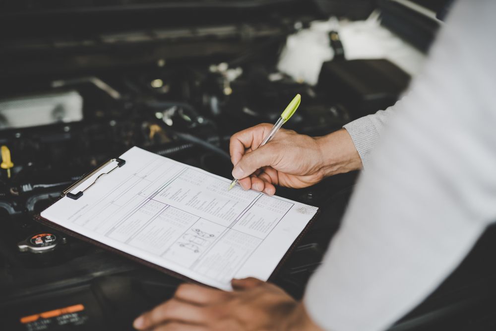 A Man Is Writing on A Clipboard in Front of A Car Engine — Rasmussen Mechanical & Tyres in Garbutt, QLD