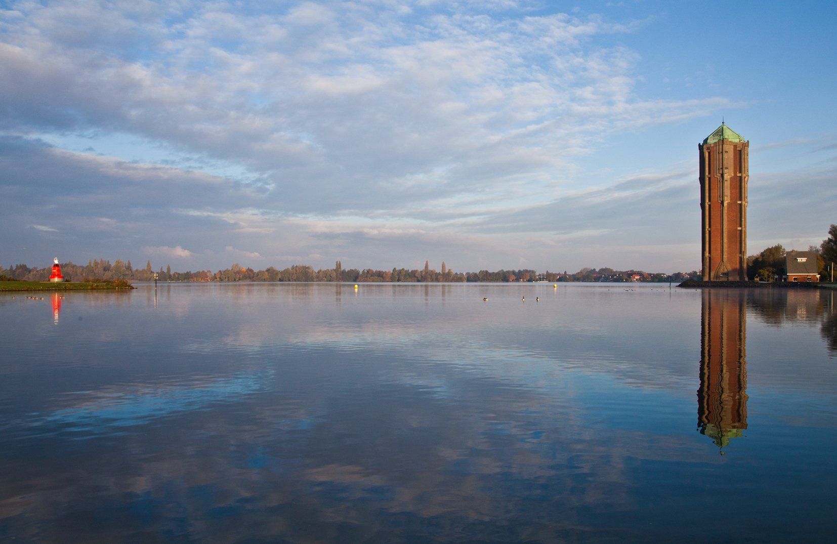 Een hoge bakstenen watertoren weerspiegelt zich in een kalm meer onder een bewolkte blauwe hemel bij zonsondergang, met de skyline van een stad in de verte.