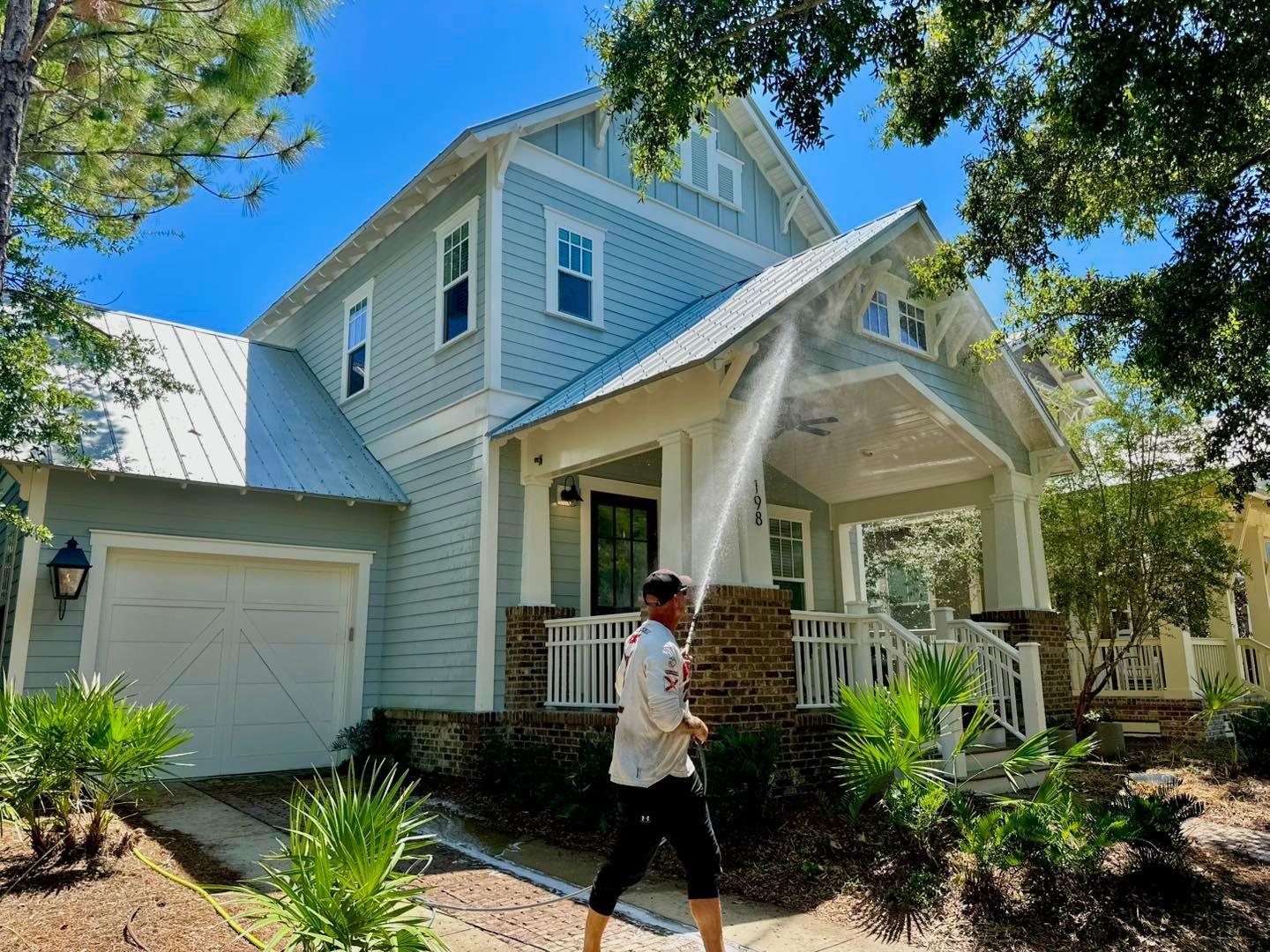 A person pressure washes the exterior of a light blue two-story house with a white garage.