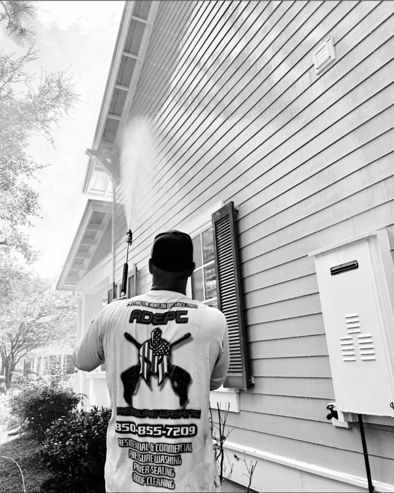 A worker in a branded t-shirt uses a power washer to clean the siding of a house.