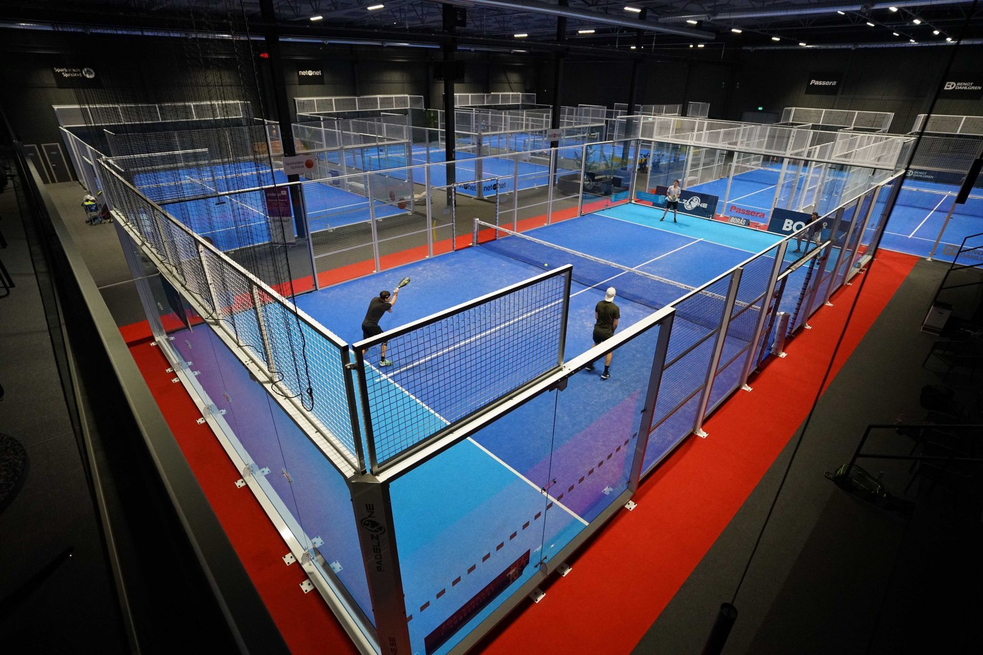 Indoor padel court with blue flooring, red borders, glass walls, and players near the net