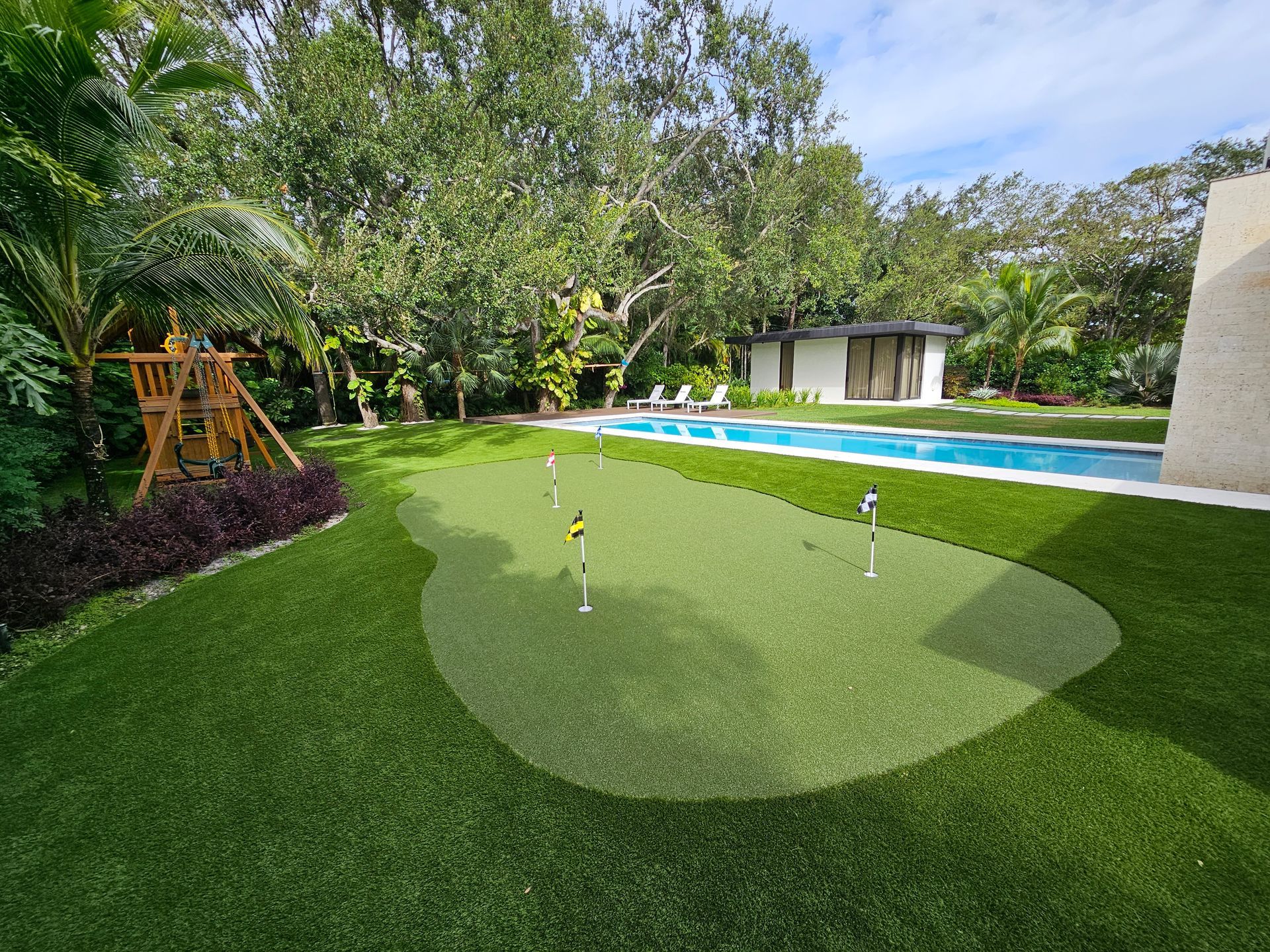 Backyard putting green with a golf hole, blue pool, and lush trees beside a modern home