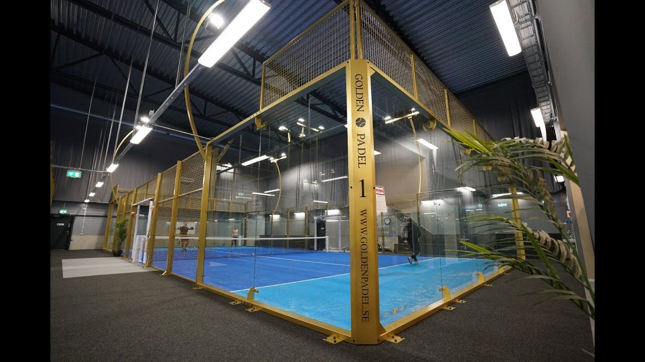 Indoor golf practice cage with yellow frame and netting over blue turf under bright lights