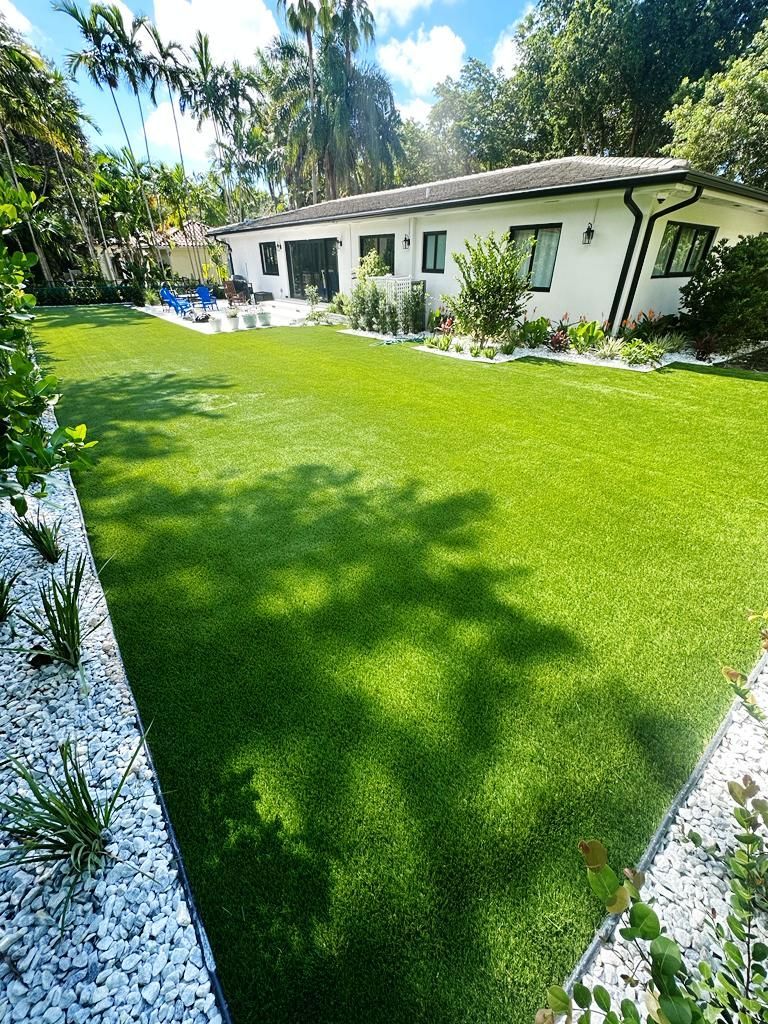 Sunny backyard with a lush green lawn, white house, palm trees, and flower beds along the edges