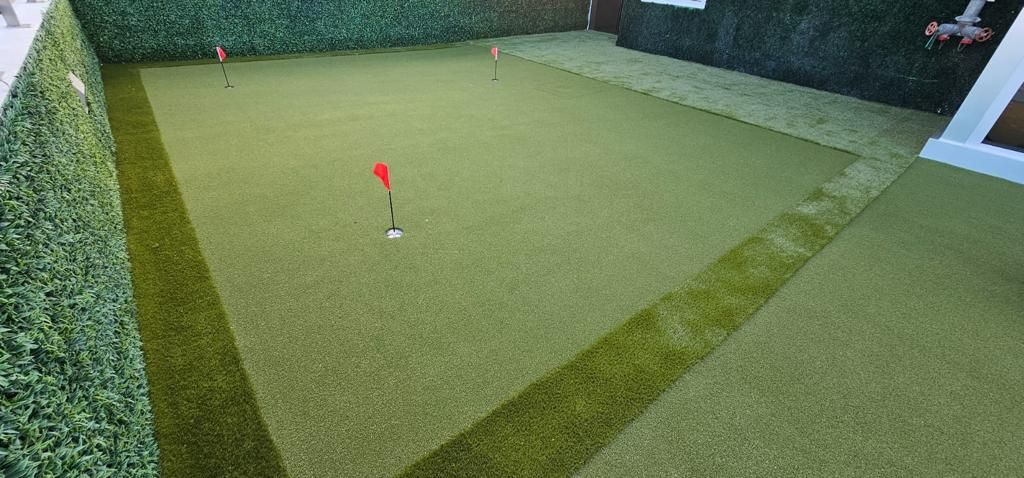Small artificial grass putting green in a walled backyard, with a red flag and golf ball near center.