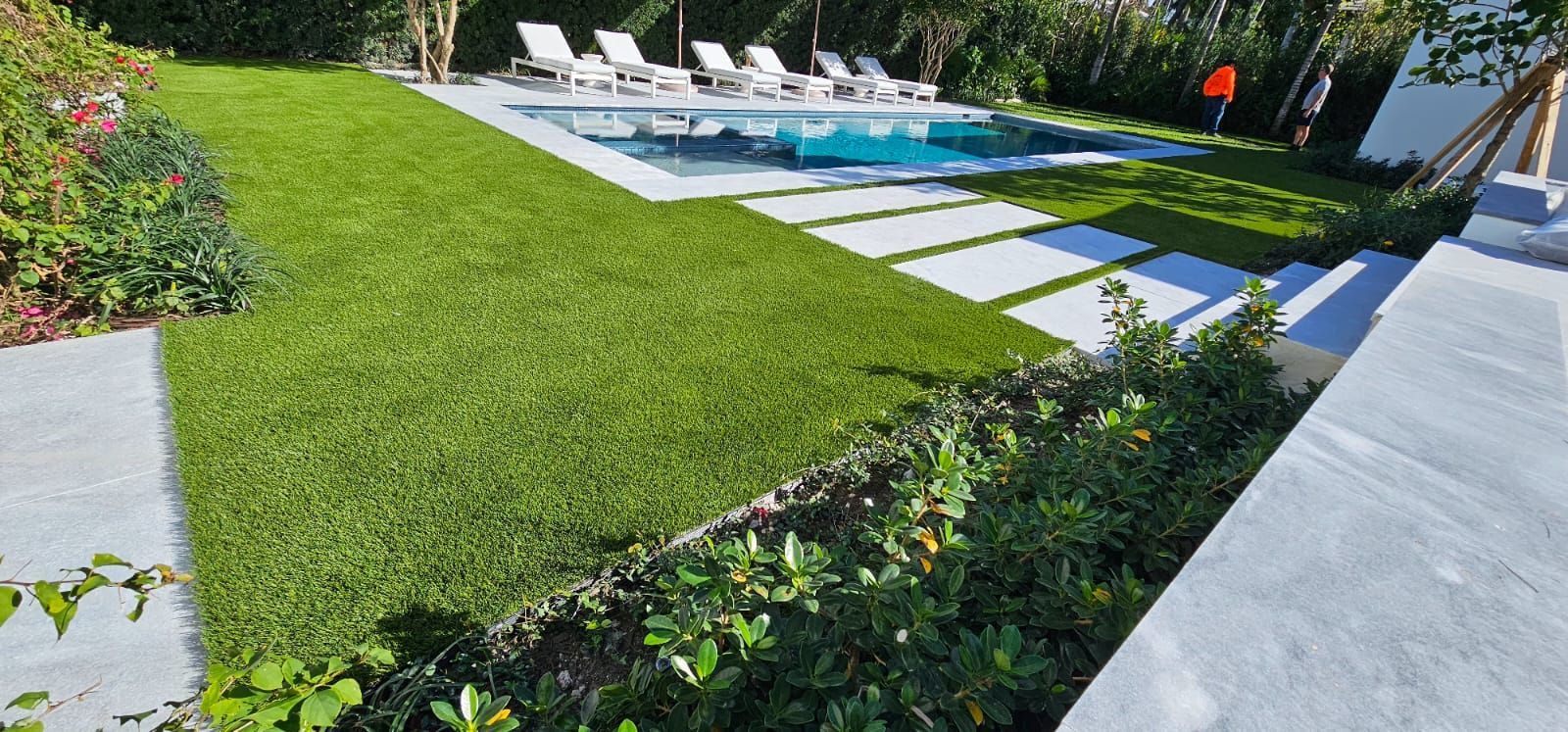 Resort pool with white loungers, blue water, and manicured green lawn by a hedge-lined walkway