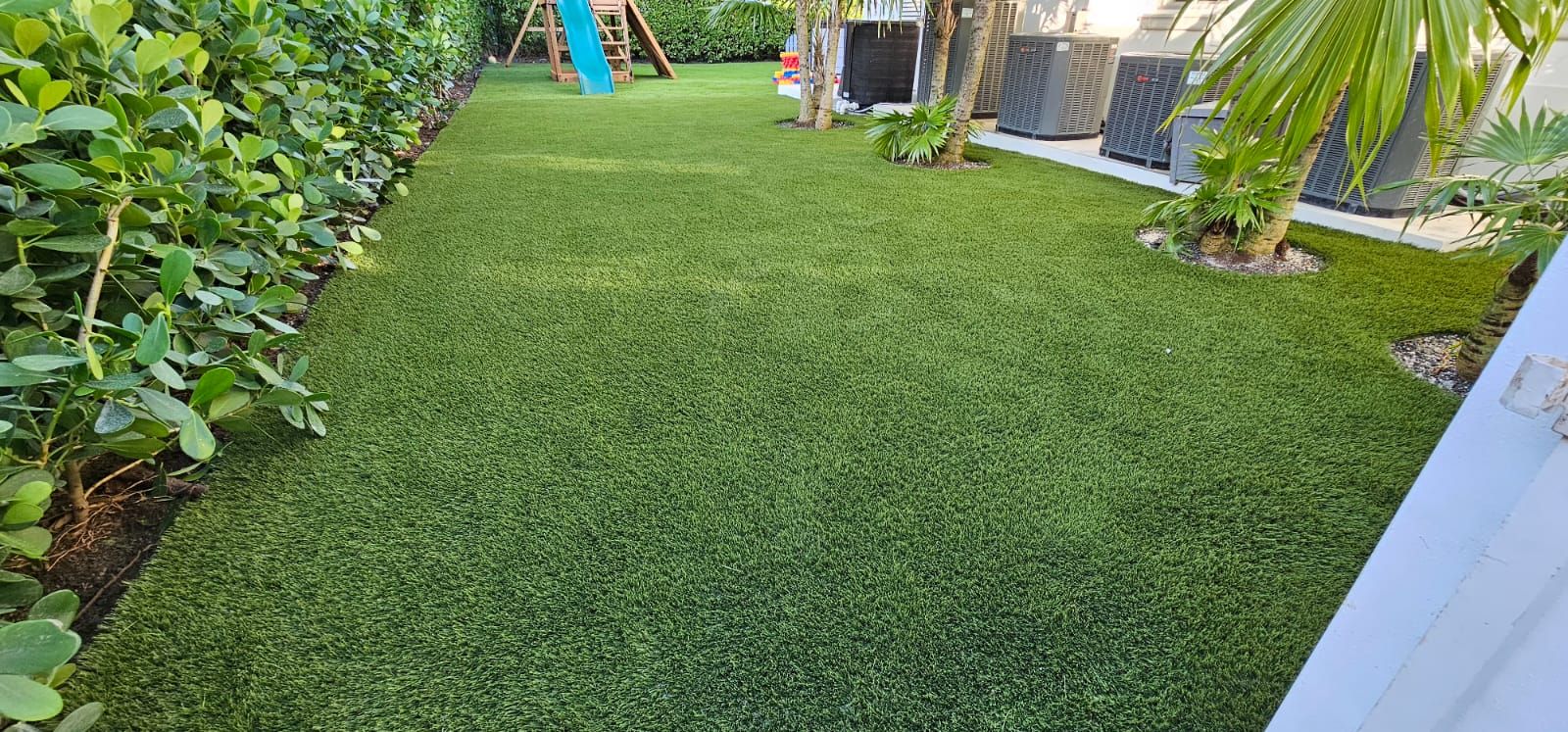 Green backyard lawn with bushes, palm trees, and a small play structure in the distance