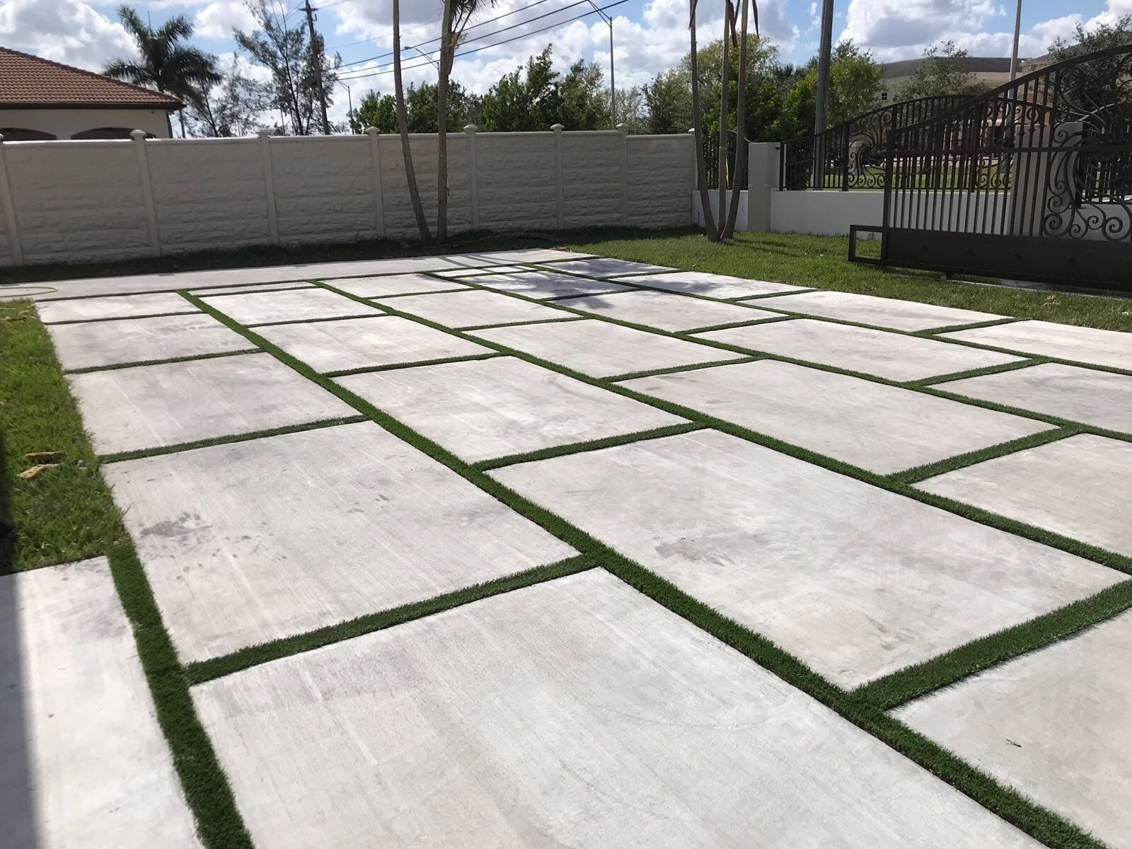 Freshly paved concrete driveway with large rectangular slabs and green grass joints, beside a fence and yard