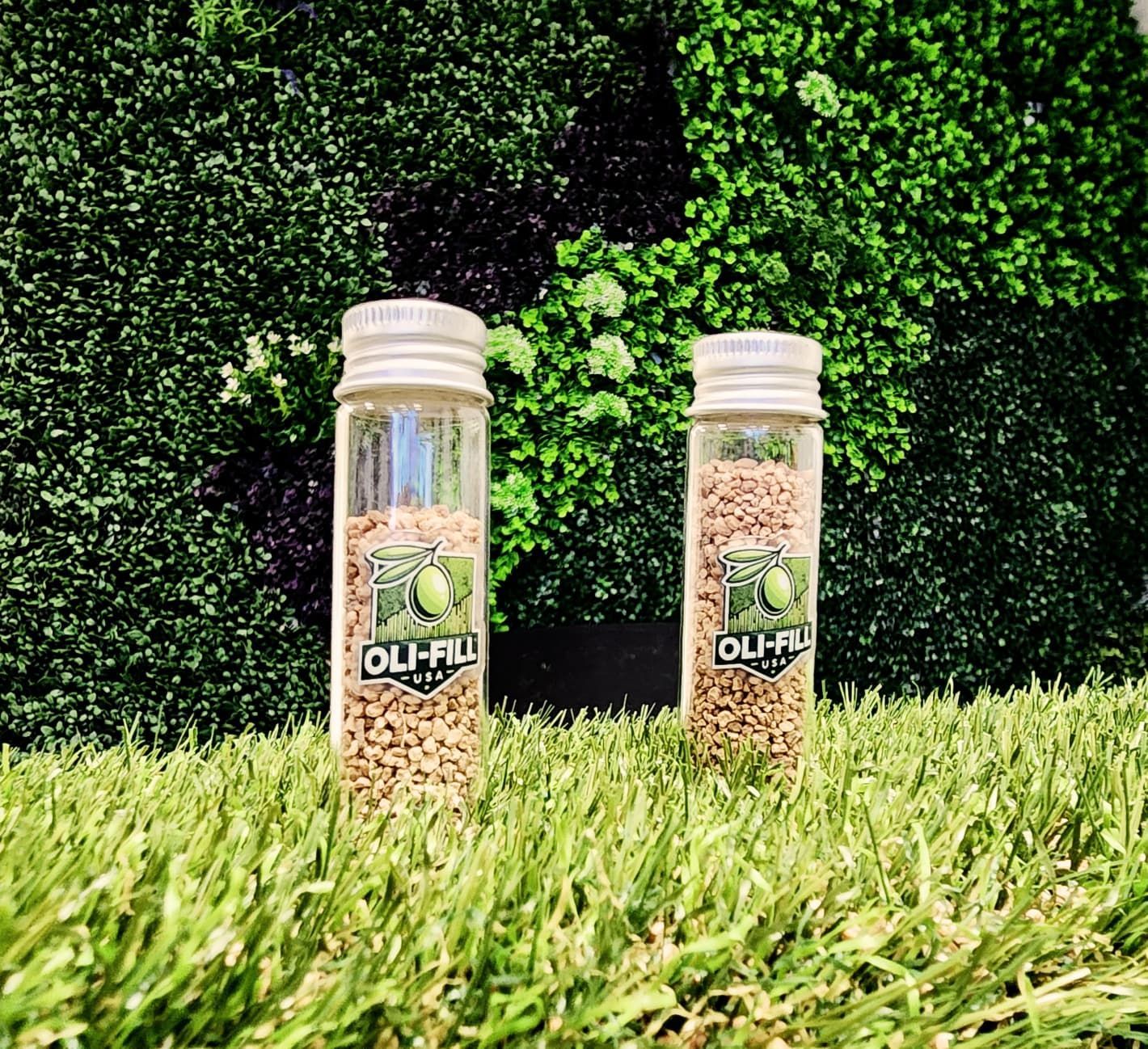 olifill sample jars Two clear glass vials with seed-like granules stand on artificial grass against a green wall; the label reads