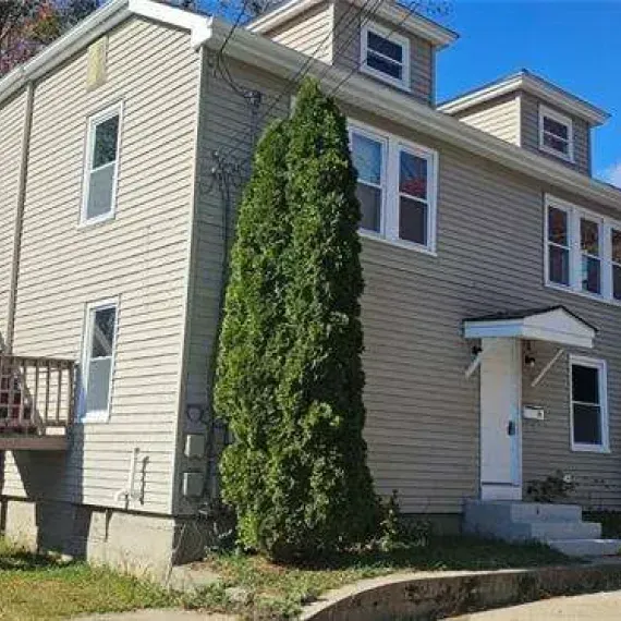 Two-story beige house with white trim and tall evergreen shrub by the front entrance