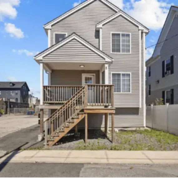 Two-story beige house with front porch, wooden stairs, and vinyl siding on a suburban street