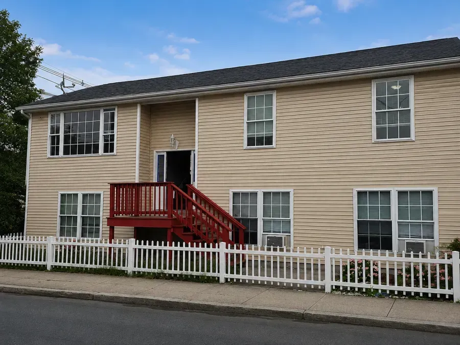 Two-story beige house with red stairs and white fence along a street