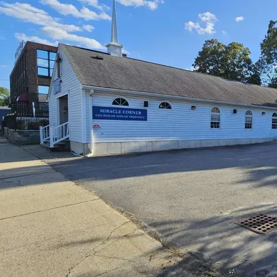 White church with steeple and blue sign on a sunny street, beside a brick building.