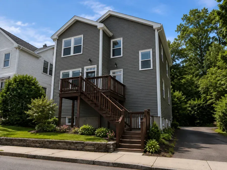 Gray house with raised wooden porch and stairs beside a driveway on a sunny street