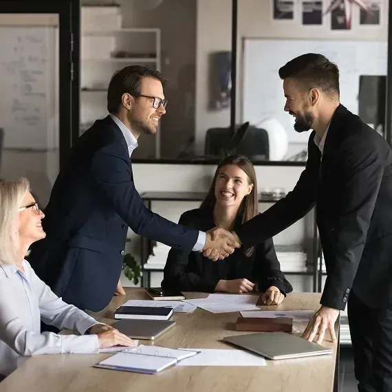 Two business professionals shake hands across a conference table in a bright office.
