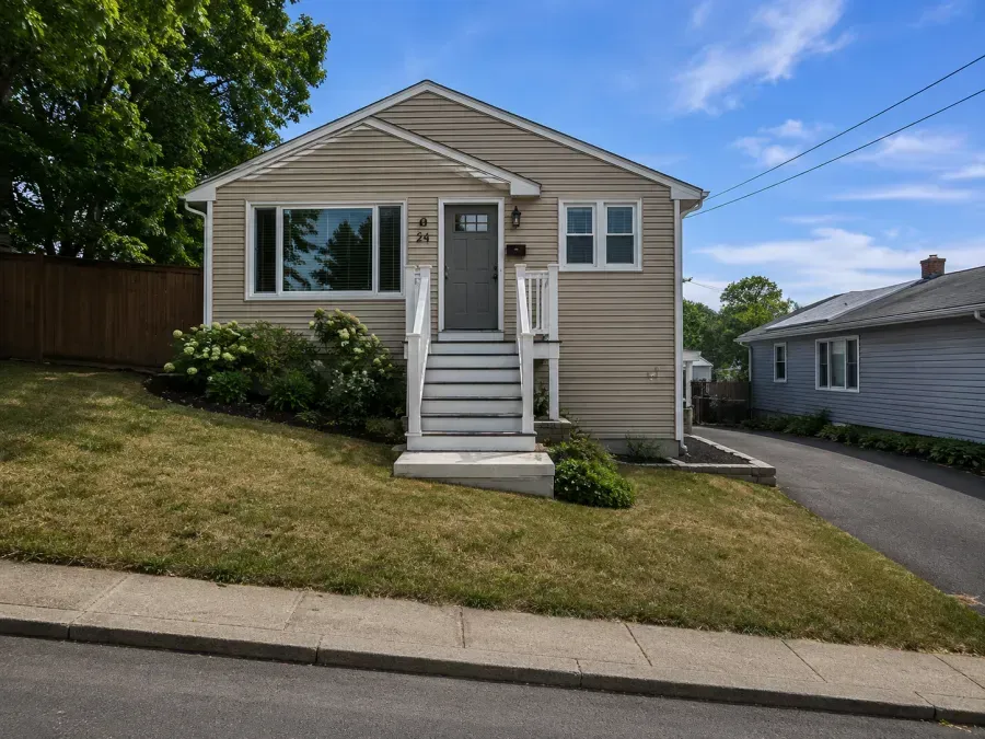 Small beige house with front steps and a sloped lawn beside a driveway on a sunny day