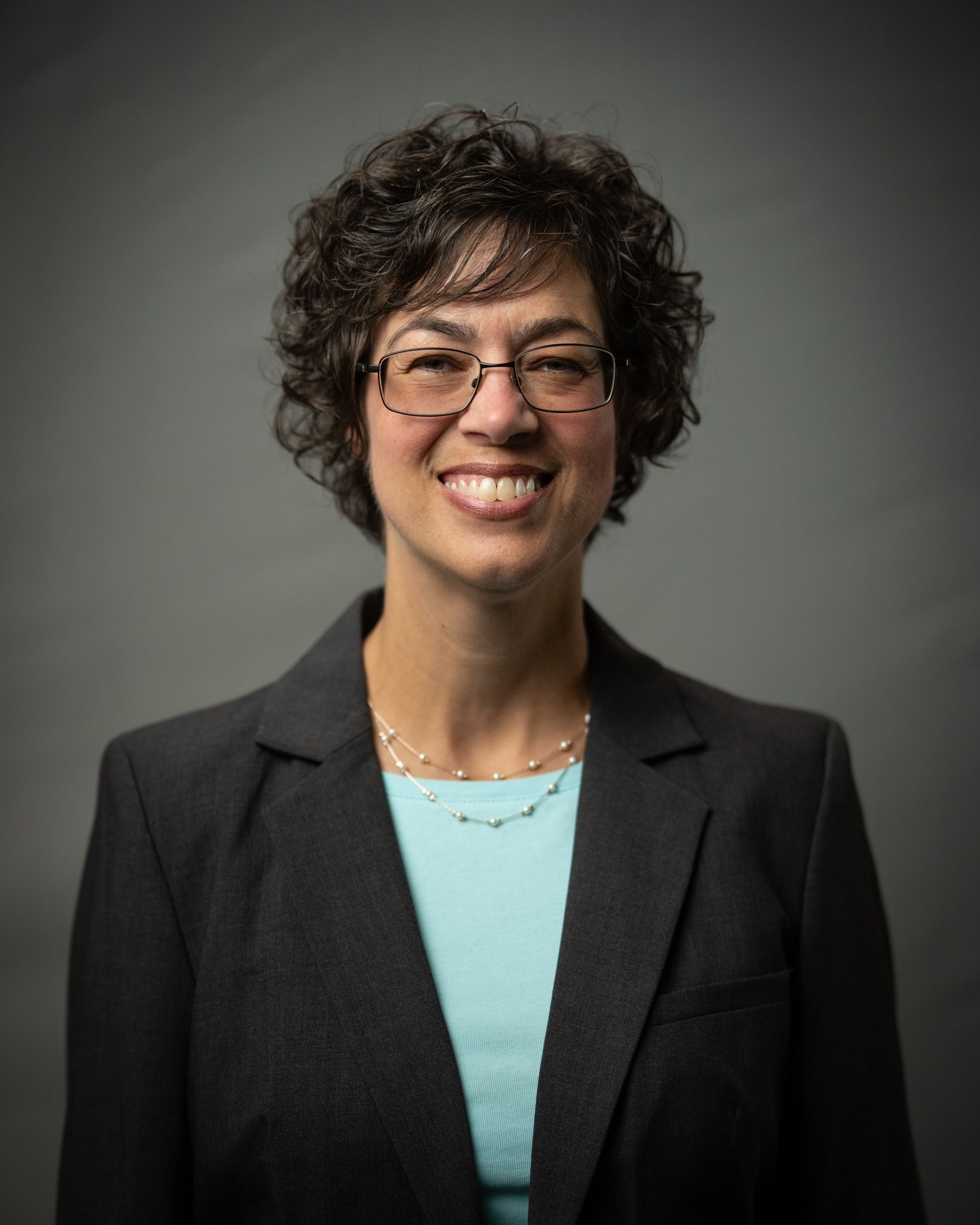 Smiling person in glasses and blazer against a dark gray studio backdrop