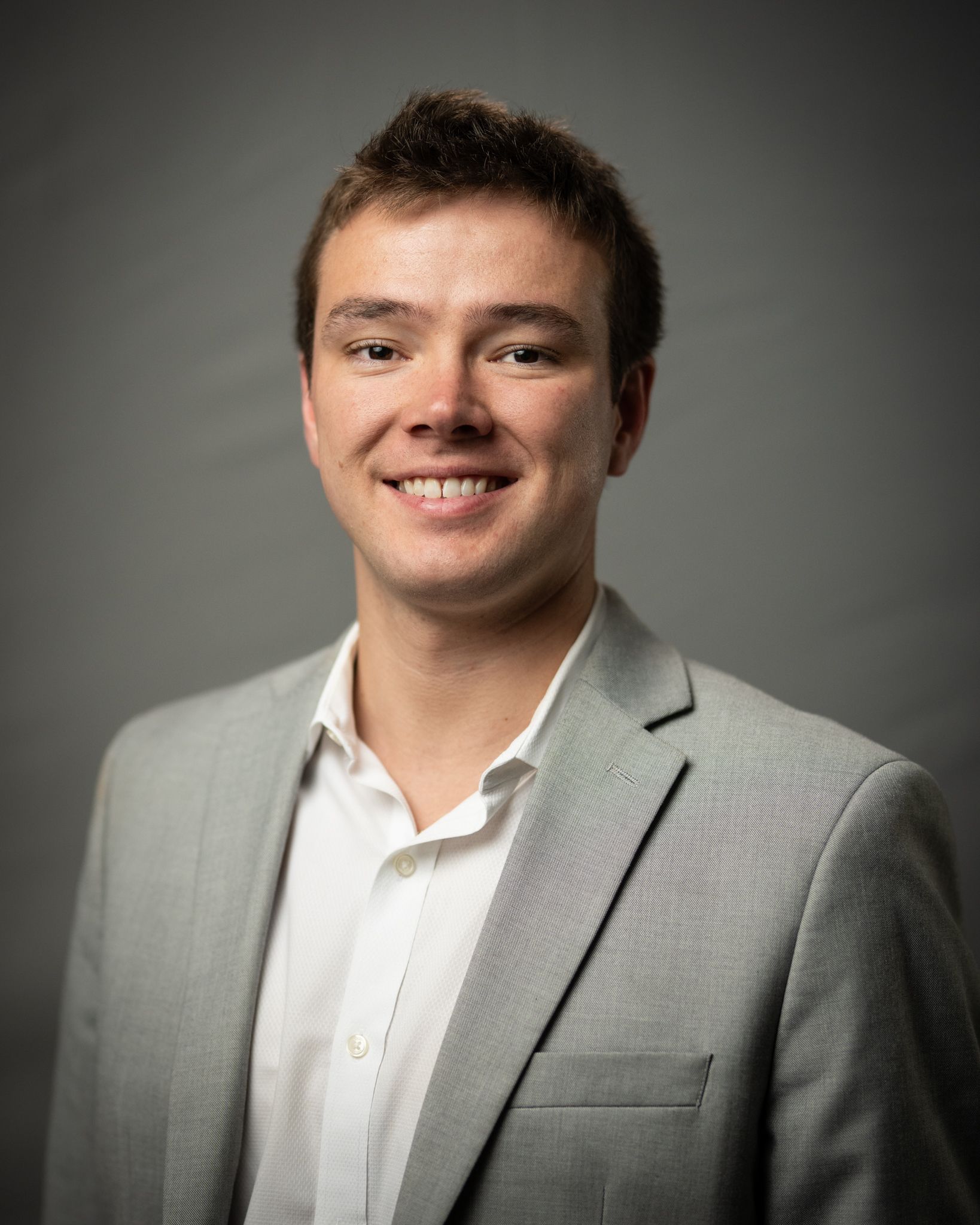 Smiling man in a light gray blazer and white shirt against a dark studio background