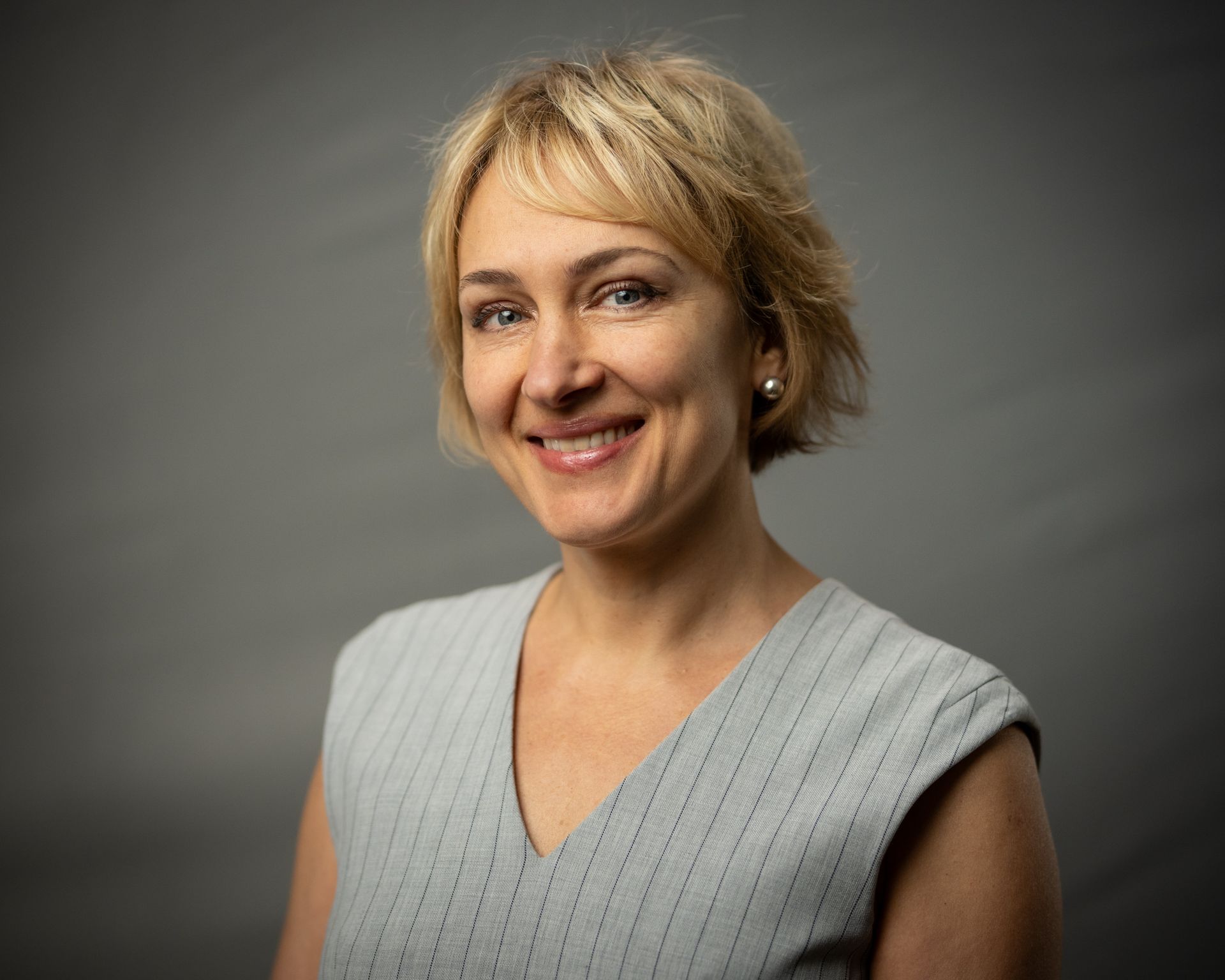 Smiling blonde woman in a light gray sleeveless top against a dark gray background