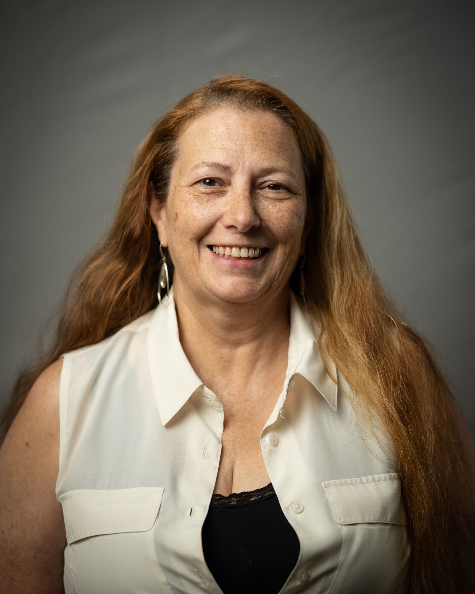 Smiling woman with long hair in a white sleeveless blouse against a gray background