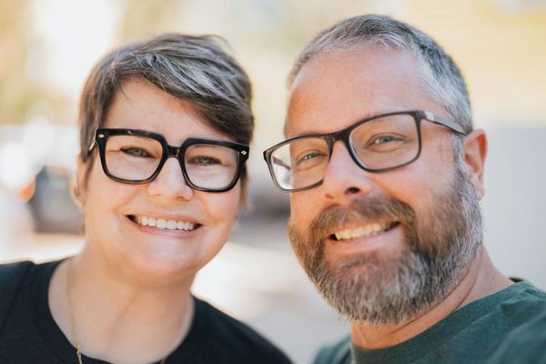 Two smiling people wearing glasses, posing closely together outdoors in soft sunlight.