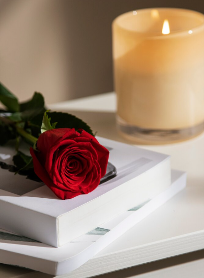 Una rosa roja sobre una pila de libros blancos junto a una vela encendida.