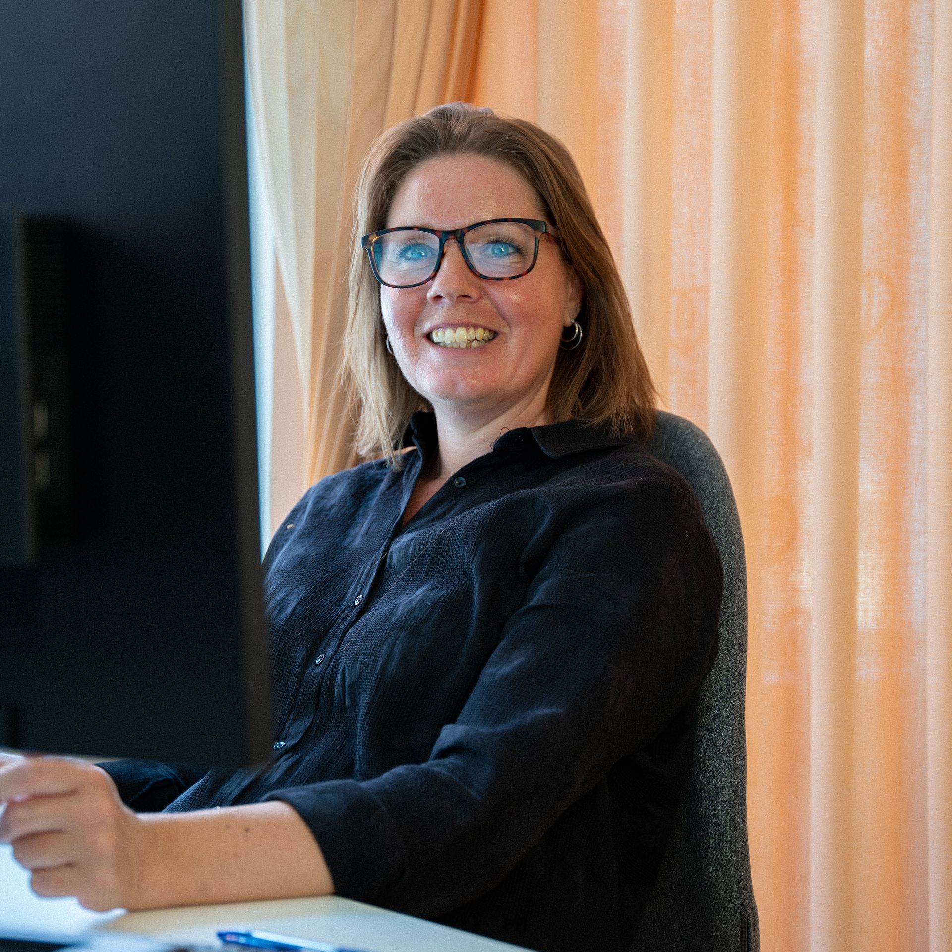 Een vrouw met een bril zit lachend achter een bureau voor een computerscherm.