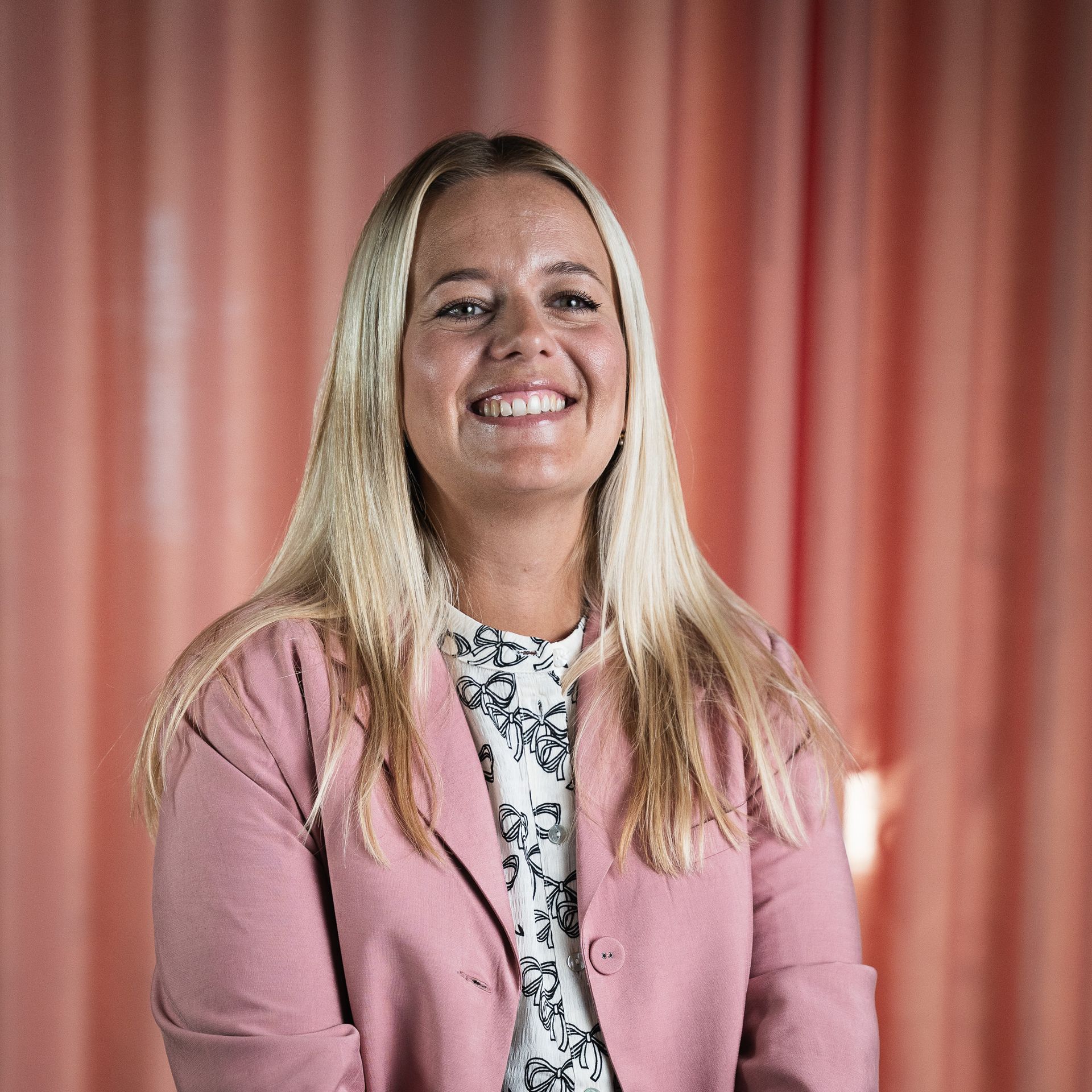 Een vrouw in een roze jasje lacht voor een roze gordijn.