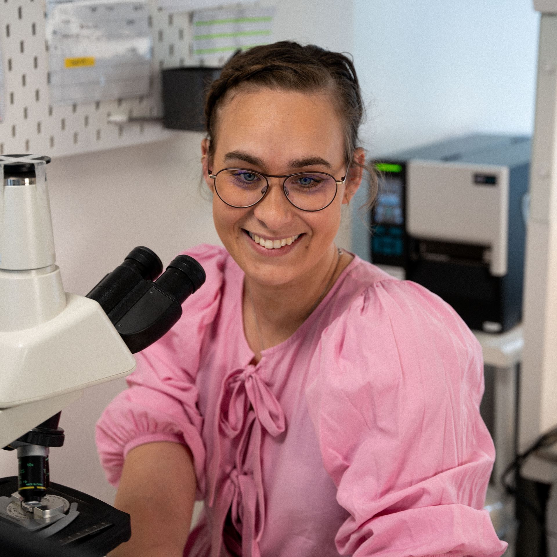Een vrouw in een roze shirt glimlacht terwijl ze door een microscoop kijkt.