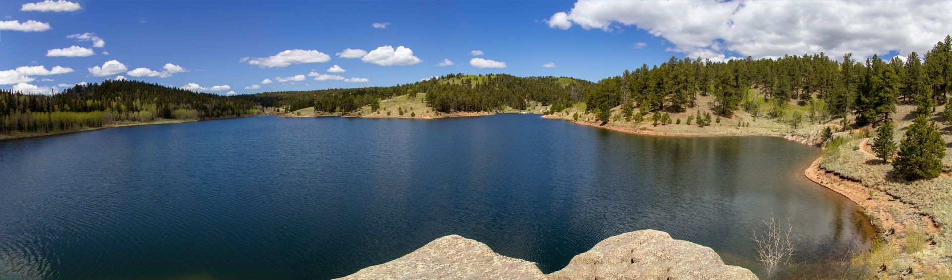Blue lake with forested shoreline, rocky foreground, and a bright sky with scattered clouds