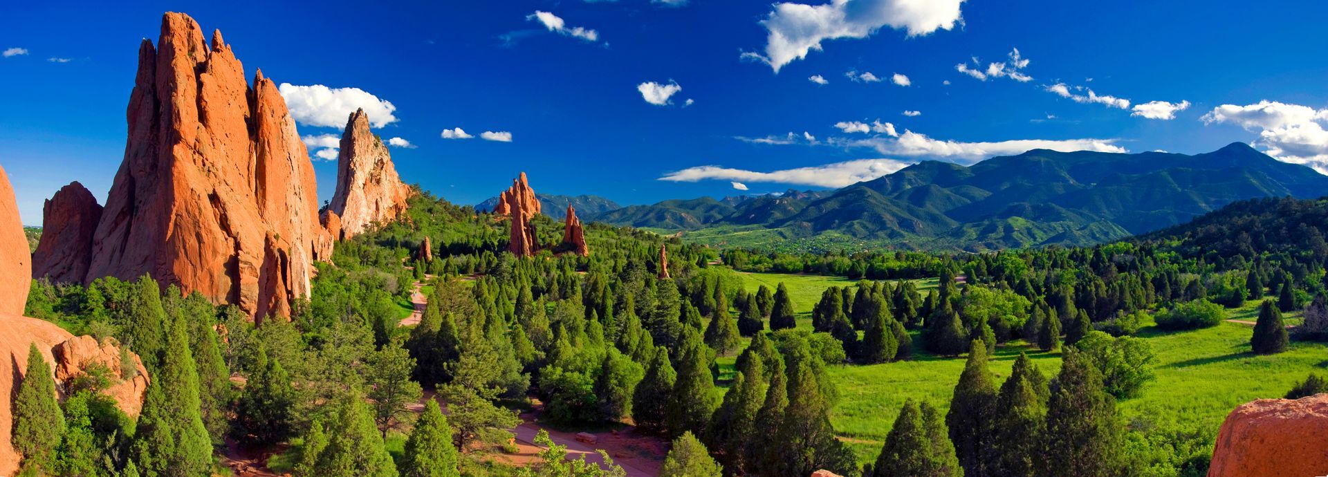 Sunlit red sandstone spires above a green valley with pine trees and mountains under a blue sky