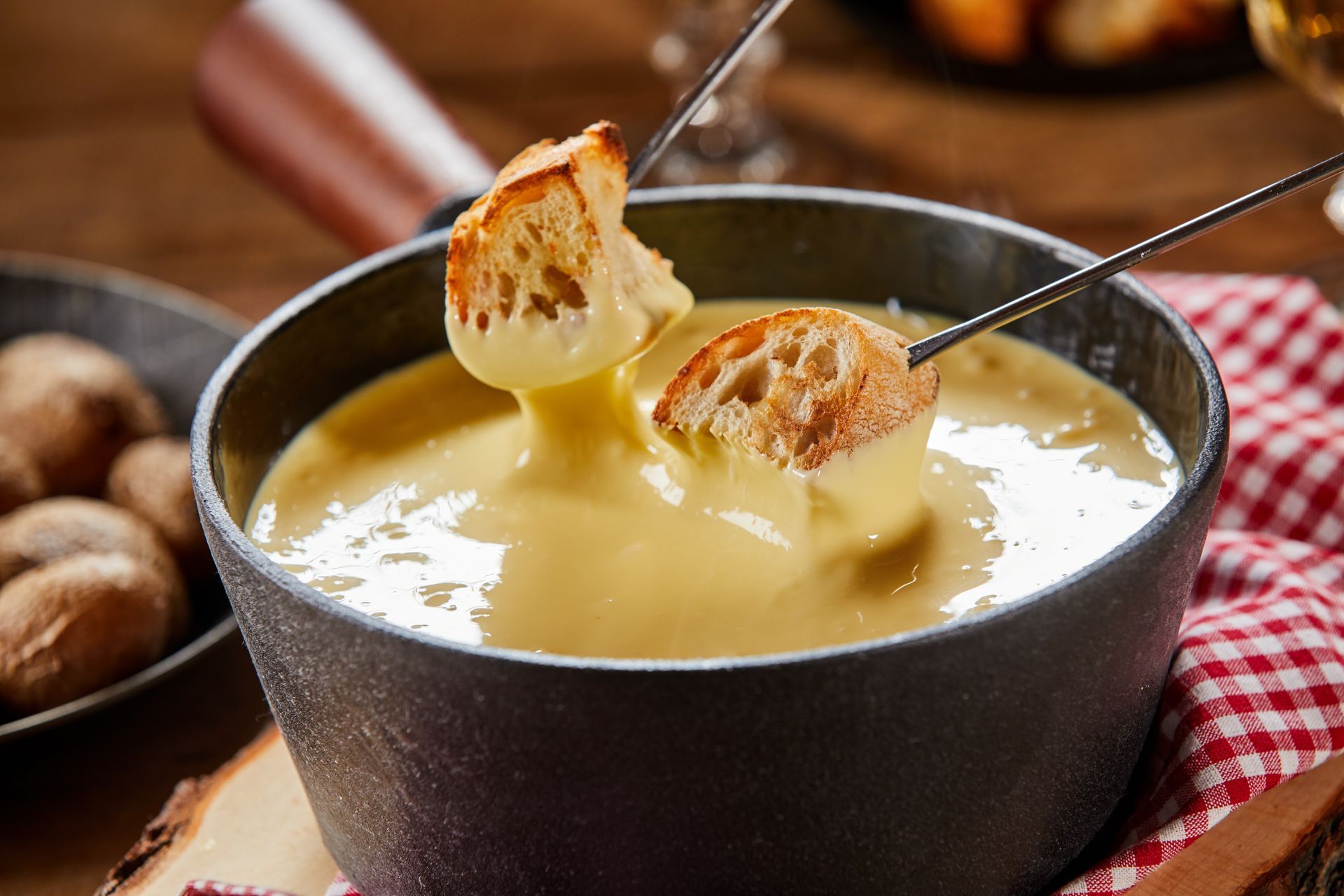 Cheese fondue in a dark pot with bread cubes on skewers, served beside potatoes on a checkered cloth