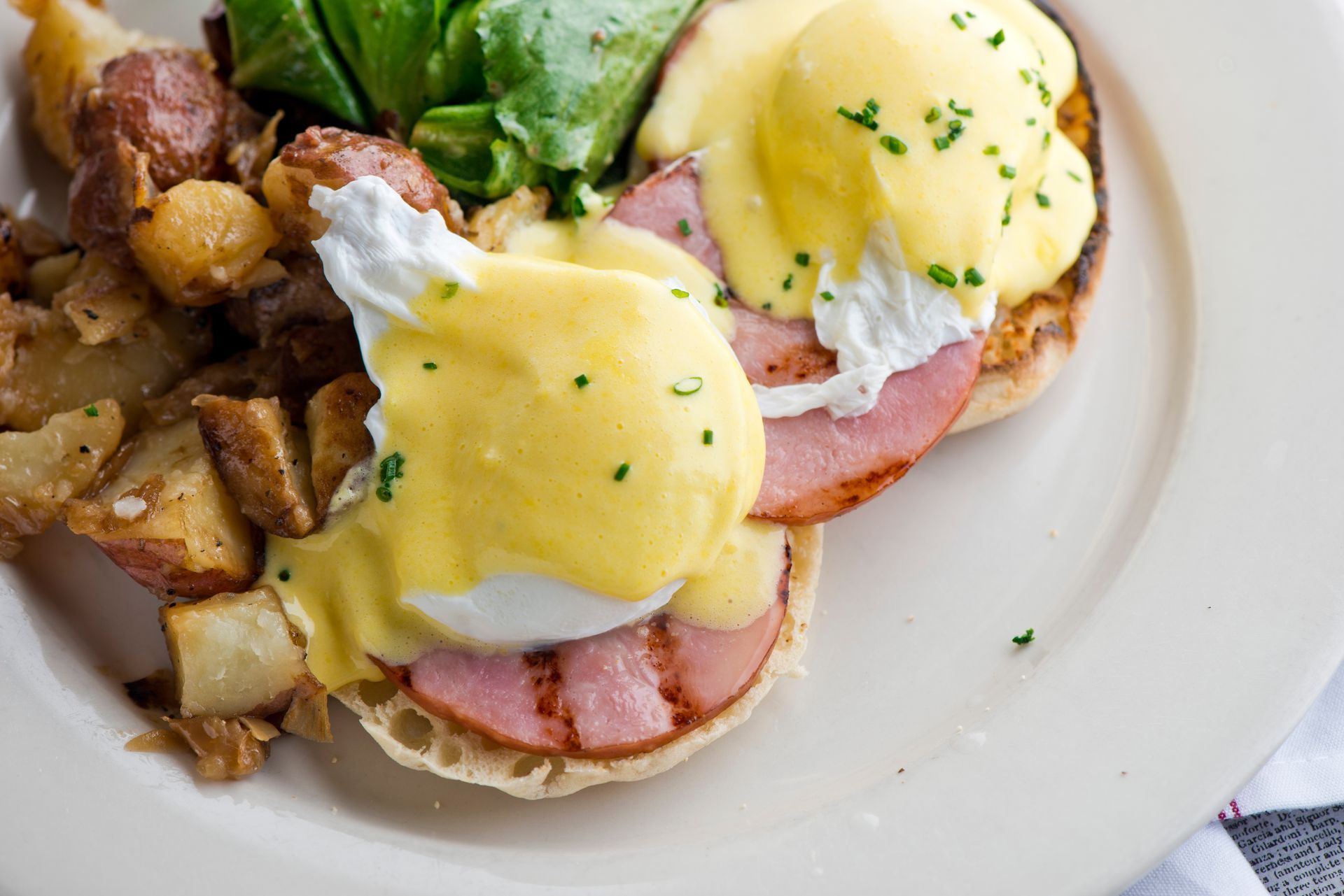 Eggs Benedict with hollandaise, ham, potatoes, and greens on a white plate.