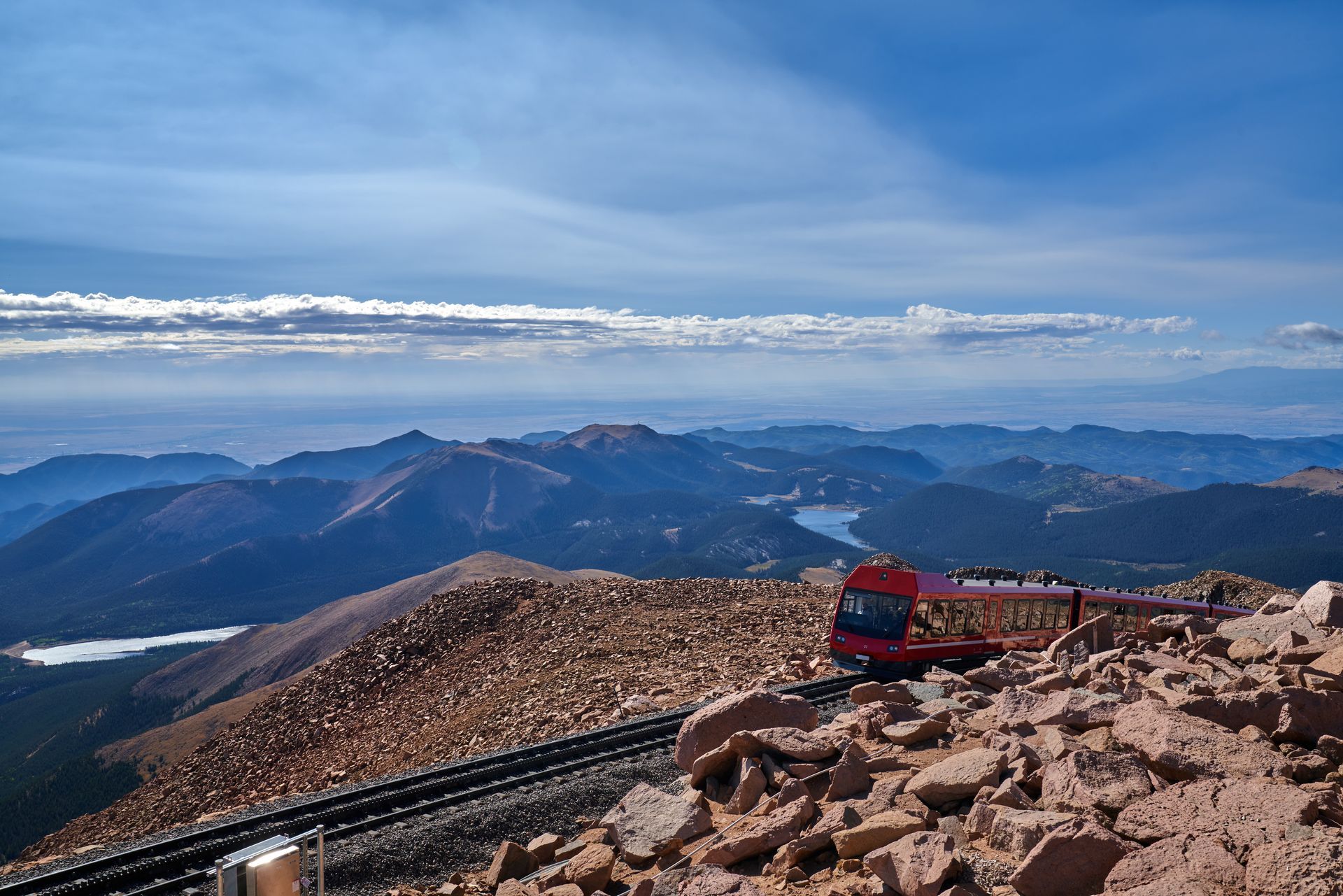 Mountain summit overlook with red train on rocky foreground and distant blue ridgelines under clear sky
