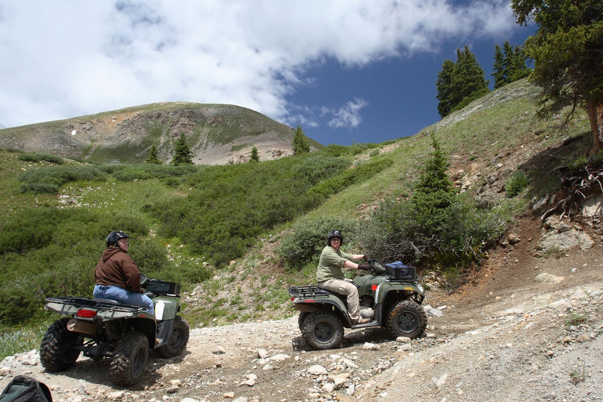 Two people riding ATVs on a rocky mountain trail under a blue sky