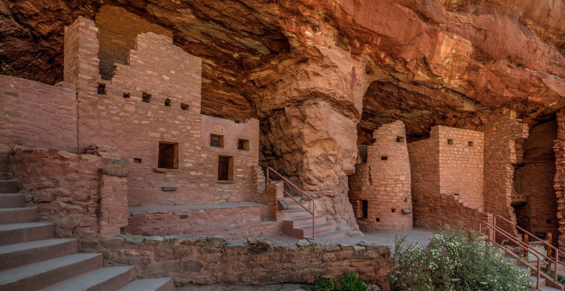 Ancient cliff dwellings carved into red sandstone beneath a rocky overhang.