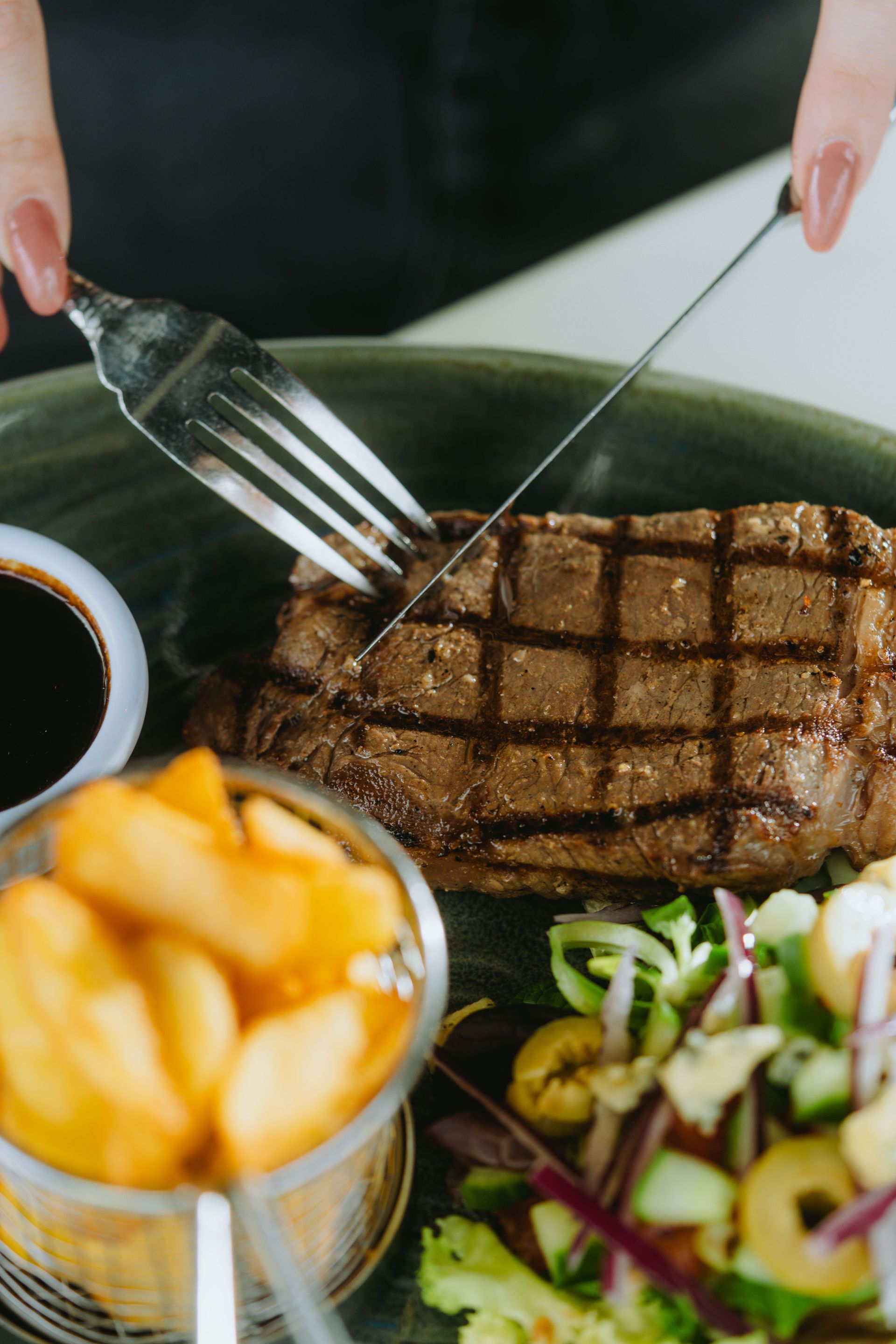Hands cutting grilled steak on a plate with fries, salad, and sauce