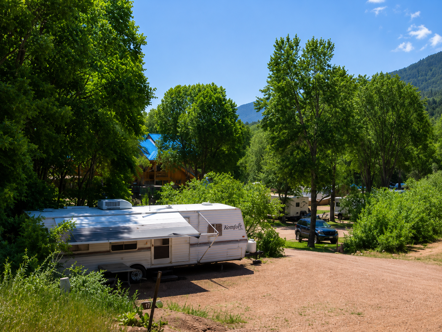 RV parked in a sunny wooded campsite with trees, a gravel lot, and mountains in the background