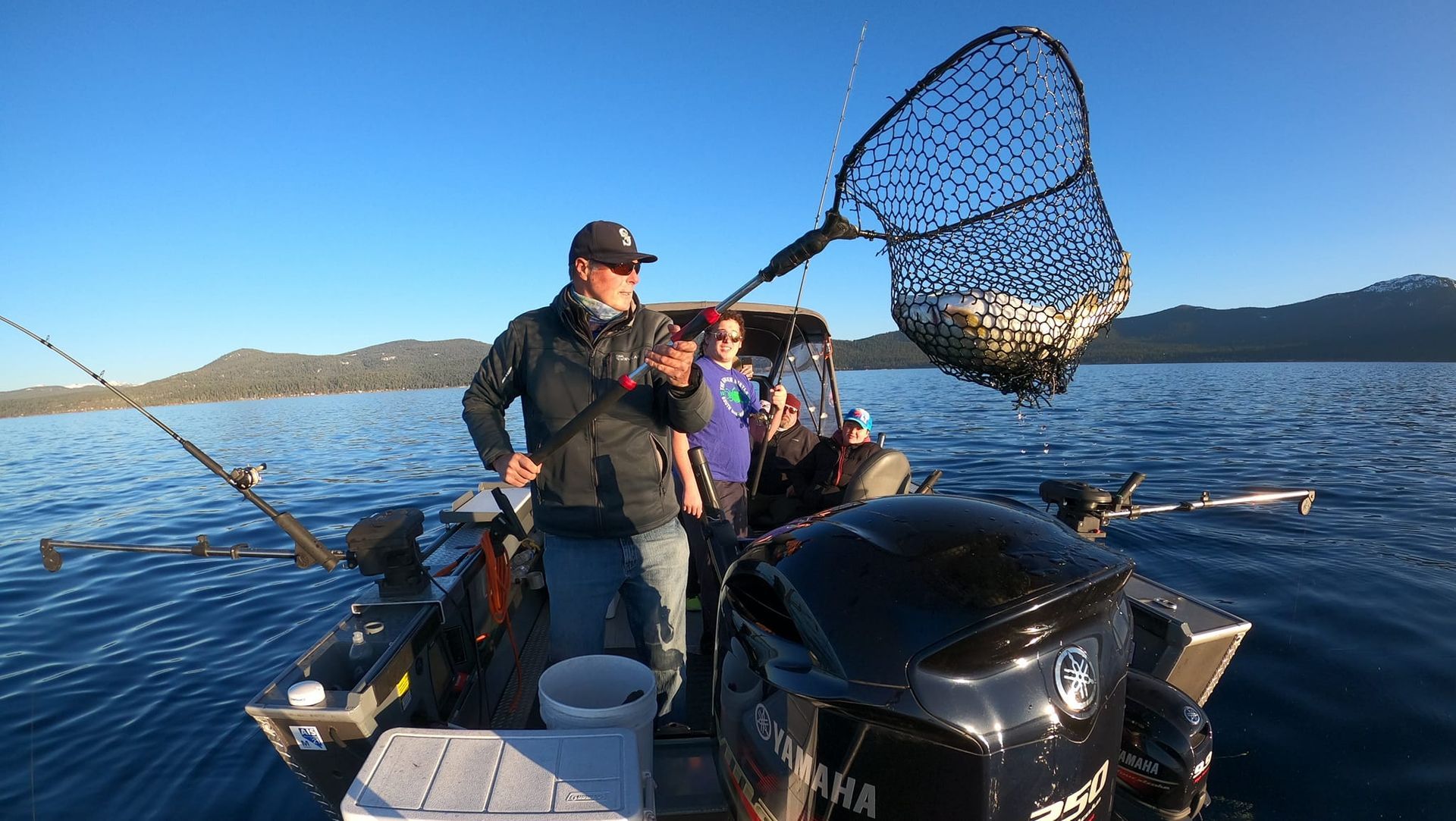 Two anglers on a boat lift a net with a fish on a sunny lake, mountains in the background.