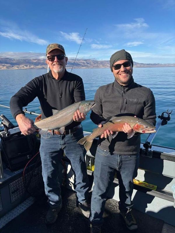 Two men on a boat holding large fish they caught, blue water and mountains in the background.