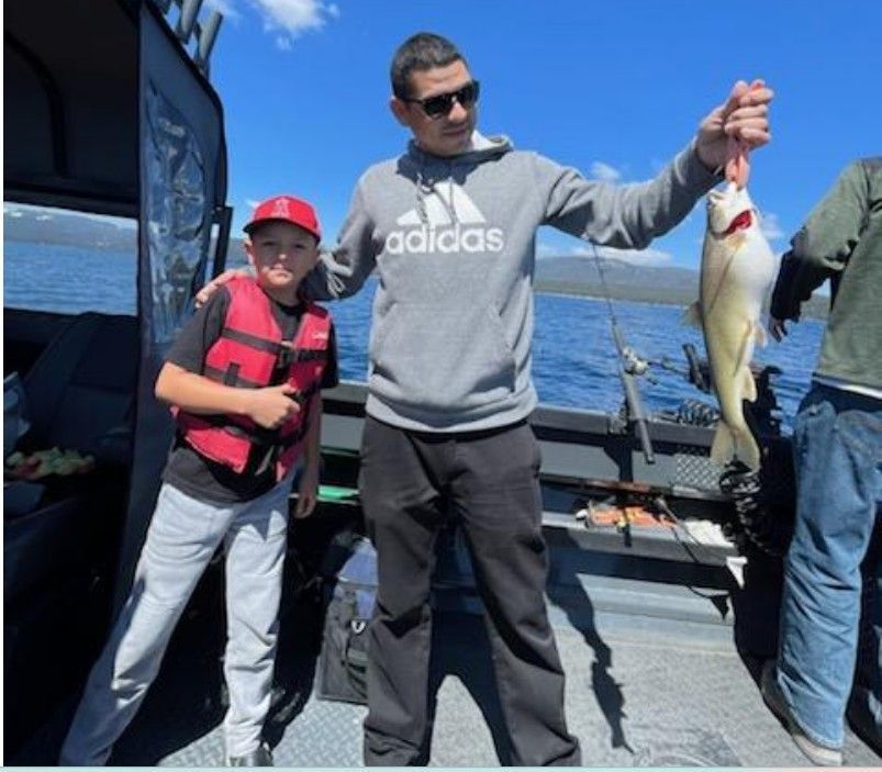 Man holding a fish on a boat; boy in a life vest looks on.